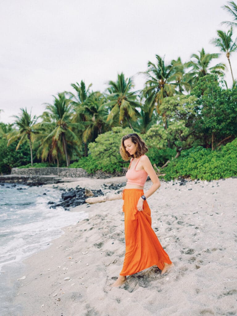 A woman in an orange skirt poses on the beach surrounded my tropical palm trees, captured by a Big Island Hawaii portrait photographer.