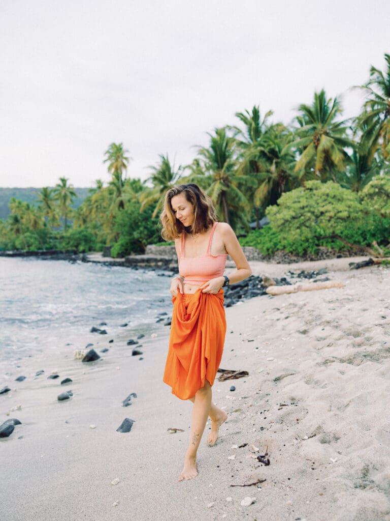 A woman in an orange skirt walking on the beach surrounded my tropical palm trees, captured by a Big Island Hawaii portrait photographer.