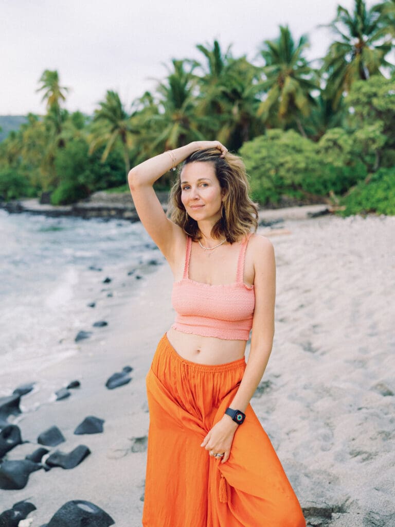 A woman in an orange skirt walking on the beach surrounded my tropical palm trees, captured by a Big Island Hawaii portrait photographer.