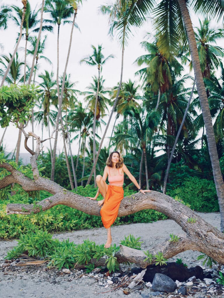 A woman in an orange skirt sitting on a palm tree on the beach surrounded my tropical nature, captured by a Big Island Hawaii portrait photographer.