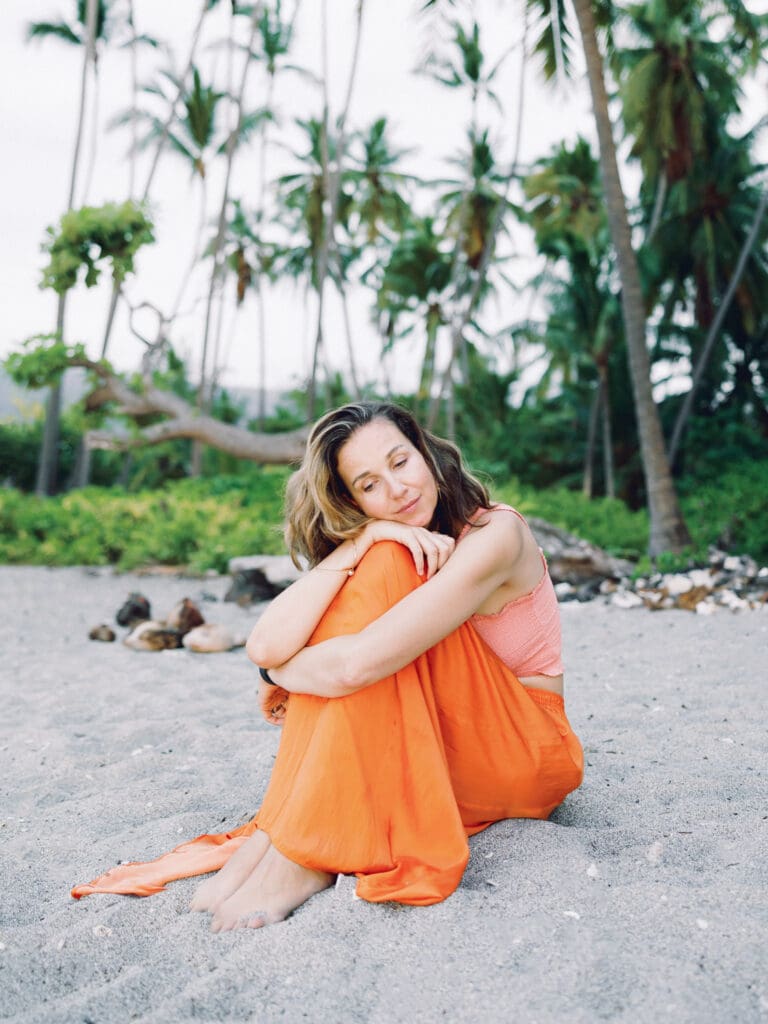 A woman in an orange skirt sitting on the beach surrounded my tropical palm trees, captured by a Big Island Hawaii portrait photographer.