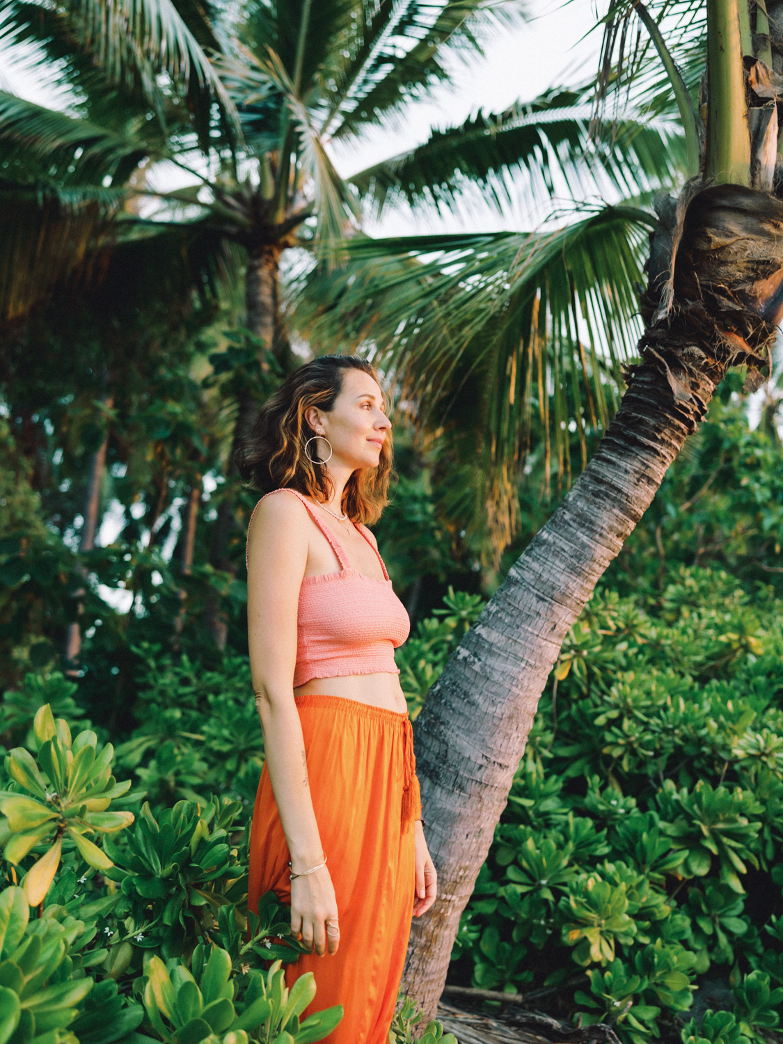 On the Big Island of Hawaii, a woman in an orange skirt stands next to a palm tree, embodying the island's tropical beauty.