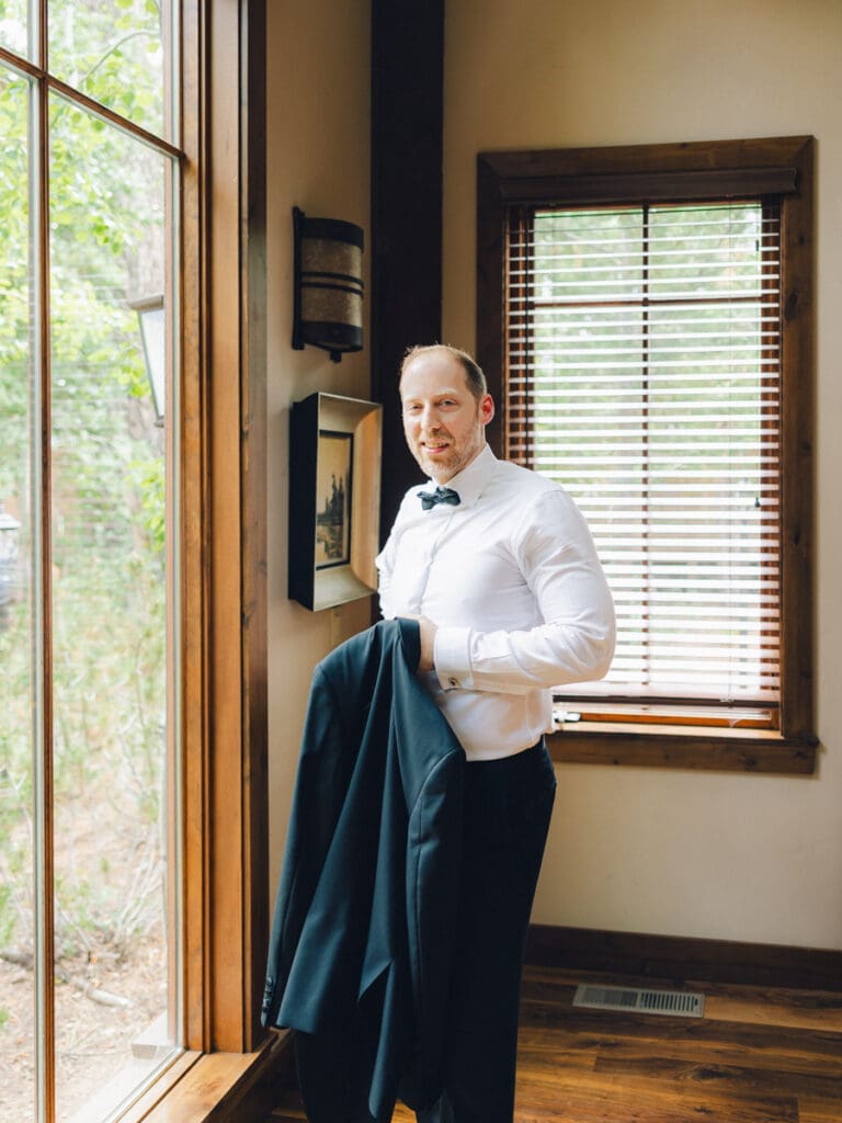 A groom in a tuxedo poses elegantly by a window at Edgewood Tahoe Resort during his wedding portrait session.