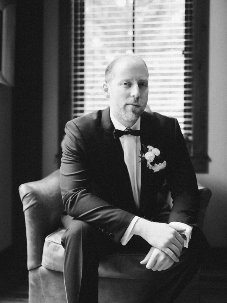 A groom in a tuxedo poses elegantly by a window at Edgewood Tahoe Resort during his wedding portrait session.