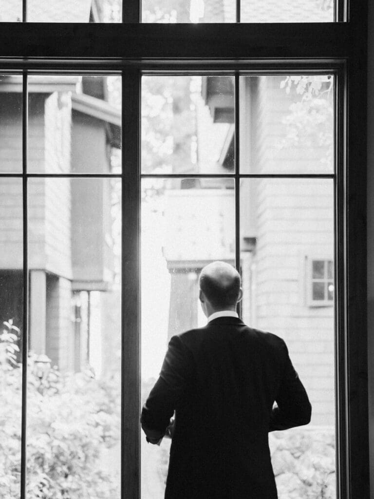 A groom in a tuxedo poses elegantly by a window at Edgewood Tahoe Resort during his wedding portrait session.