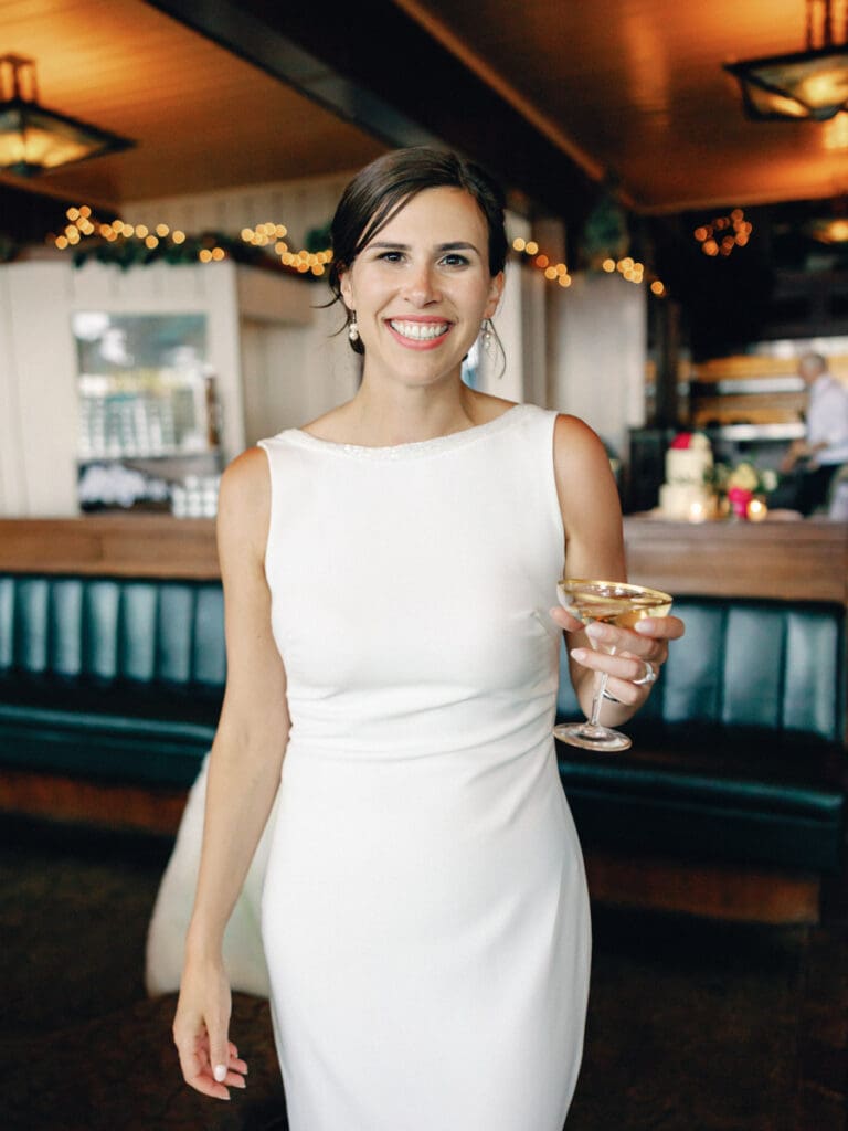 A smiling bride in a white dress holding heirloom champagne glass poses for the camera at her wedding at Gar Woods, Lake Tahoe.