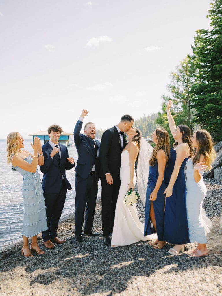 A joyful group of groomsmen and bridesmaids in blue dresses stands side by side, celebrating at a wedding at Gar Woods, Lake Tahoe.