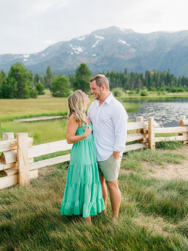 A romantic engaged couple poses by a fence, with the breathtaking Lake Tahoe mountains illuminated by a sunset backdrop.