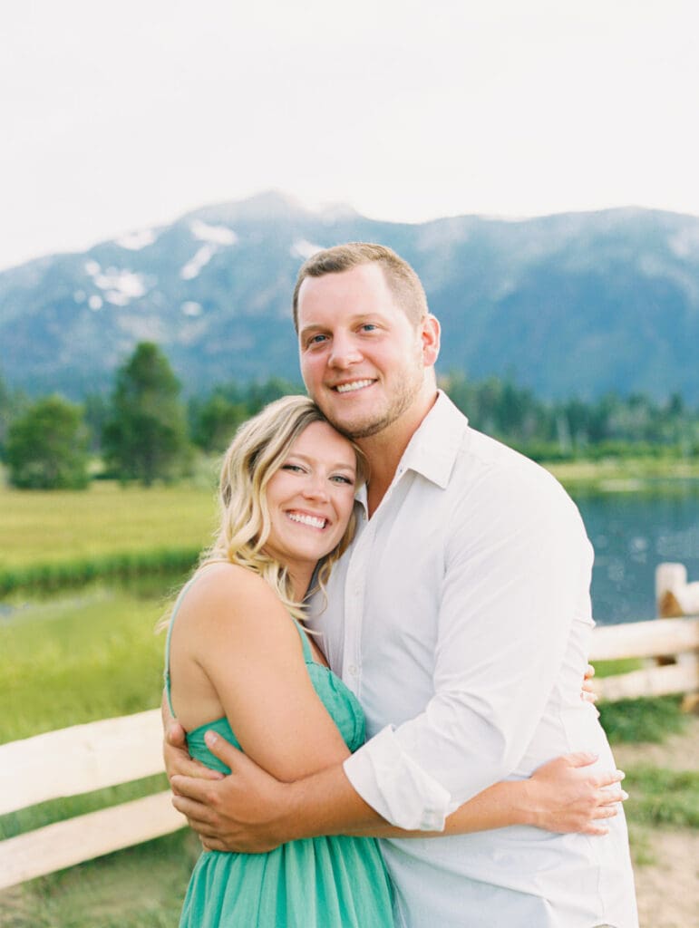 A romantic engaged couple poses by a fence, with the breathtaking Lake Tahoe mountains illuminated by a sunset backdrop.