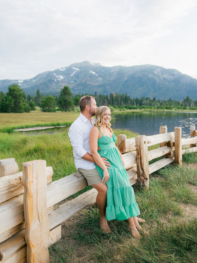 A romantic engaged couple poses by a fence, with the breathtaking Lake Tahoe mountains illuminated by a sunset backdrop.