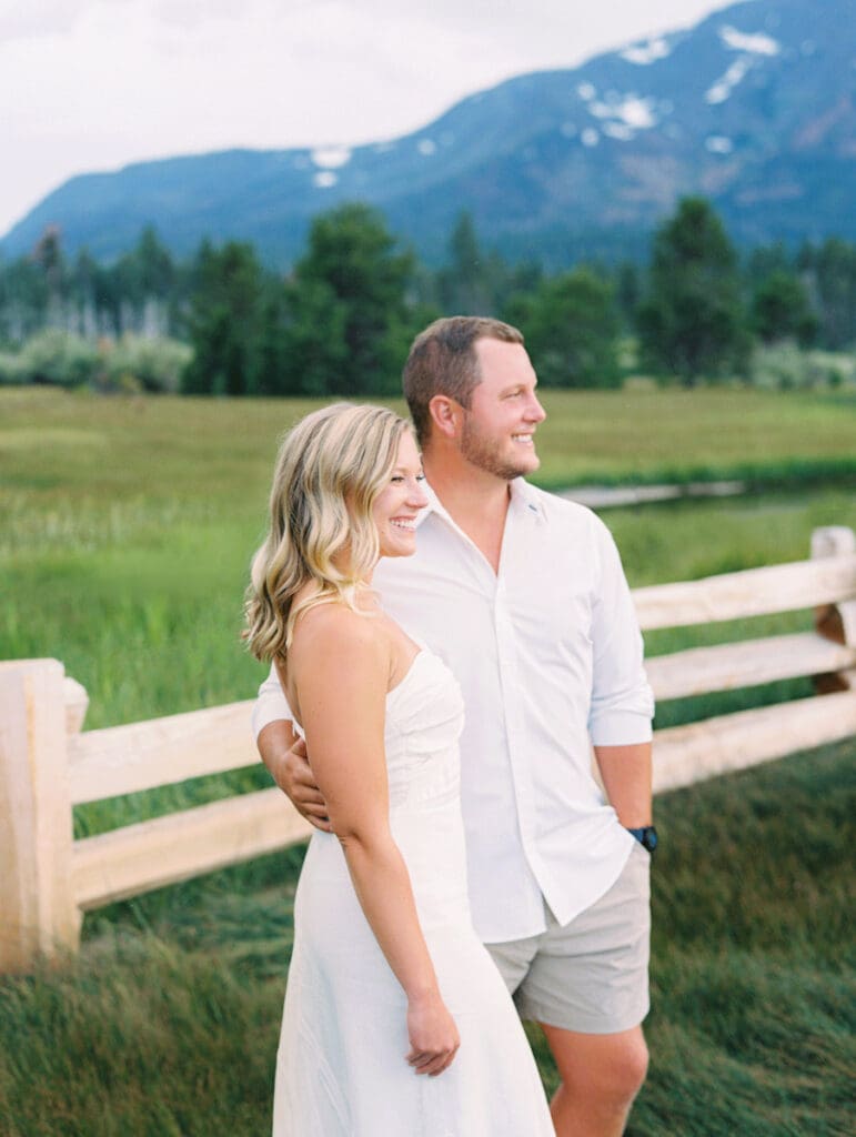 A romantic engaged couple poses by a fence, with the breathtaking Lake Tahoe mountains illuminated by a sunset backdrop.