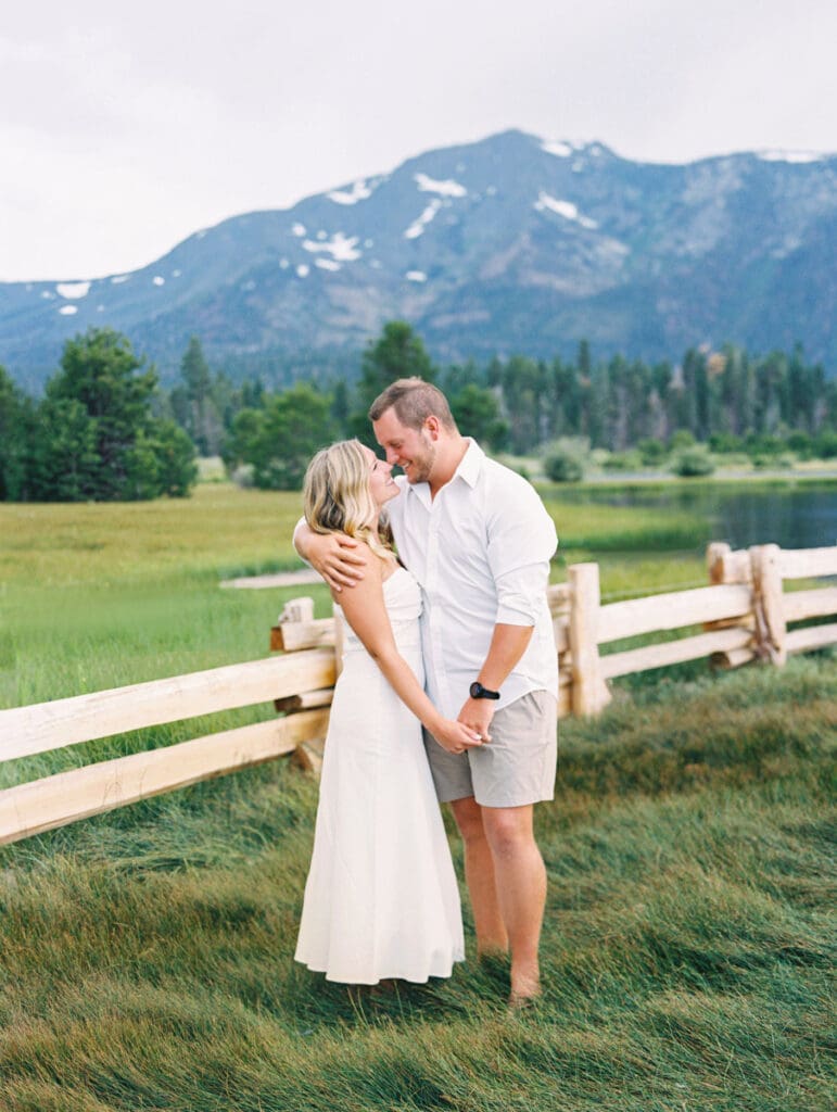 A couple smiling on the beach at Lake Tahoe, surrounded by mountains, capturing a dreamy sunset engagement moment.