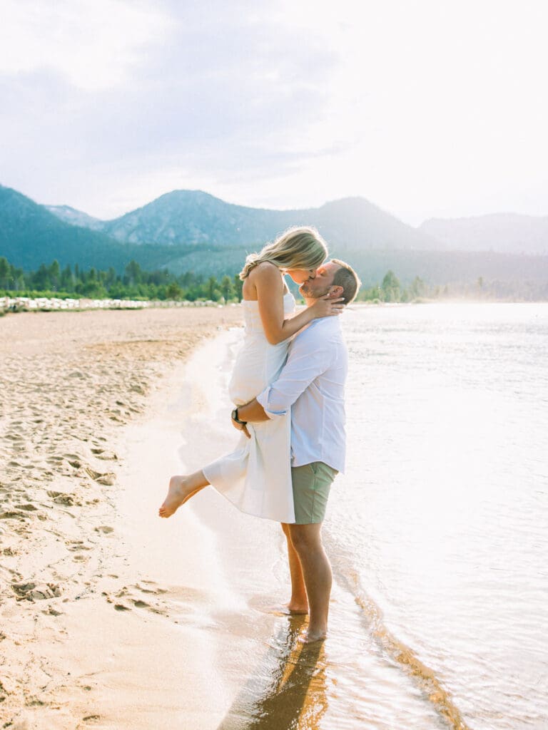 A couple kisses on the beach at Lake Tahoe, surrounded by mountains, capturing a dreamy sunset engagement moment.