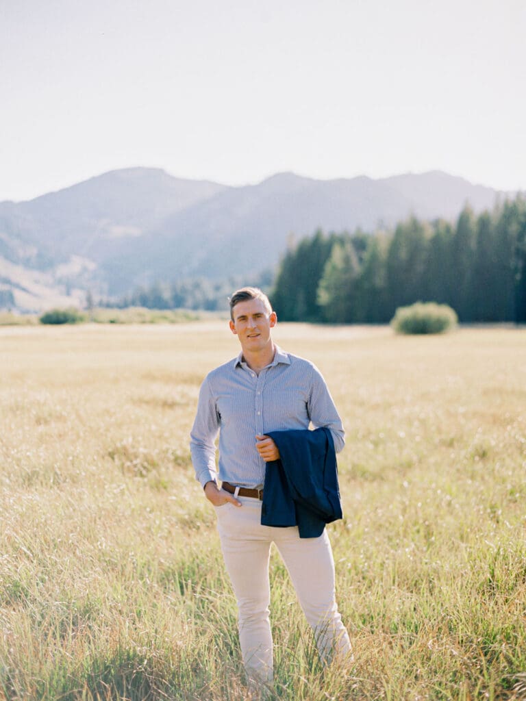 An engaged man dressed in a blue suit poses in a vibrant field, with the majestic Lake Tahoe mountains in the background.