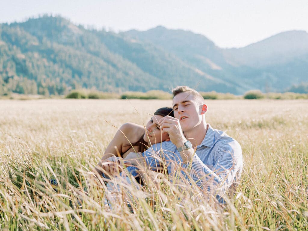 A couple is sitting in a lush alpine field, capturing their engagement amidst the majestic Lake Tahoe mountains.