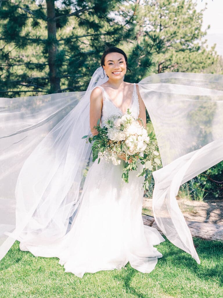 A bride in a white dress and veil stands atop a mountain, overlooking Tannenbaum Lake Tahoe during her wedding.