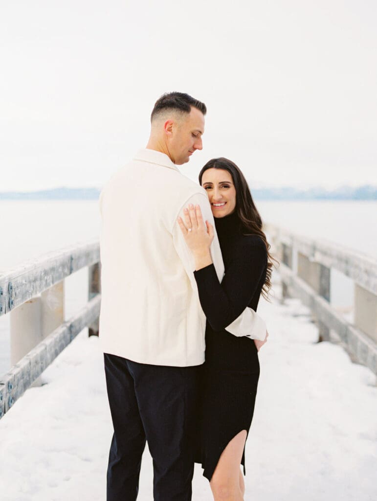 A couple stands together on a dock at Lake Tahoe, surrounded by serene water and majestic mountains in the background celebrating their engagement