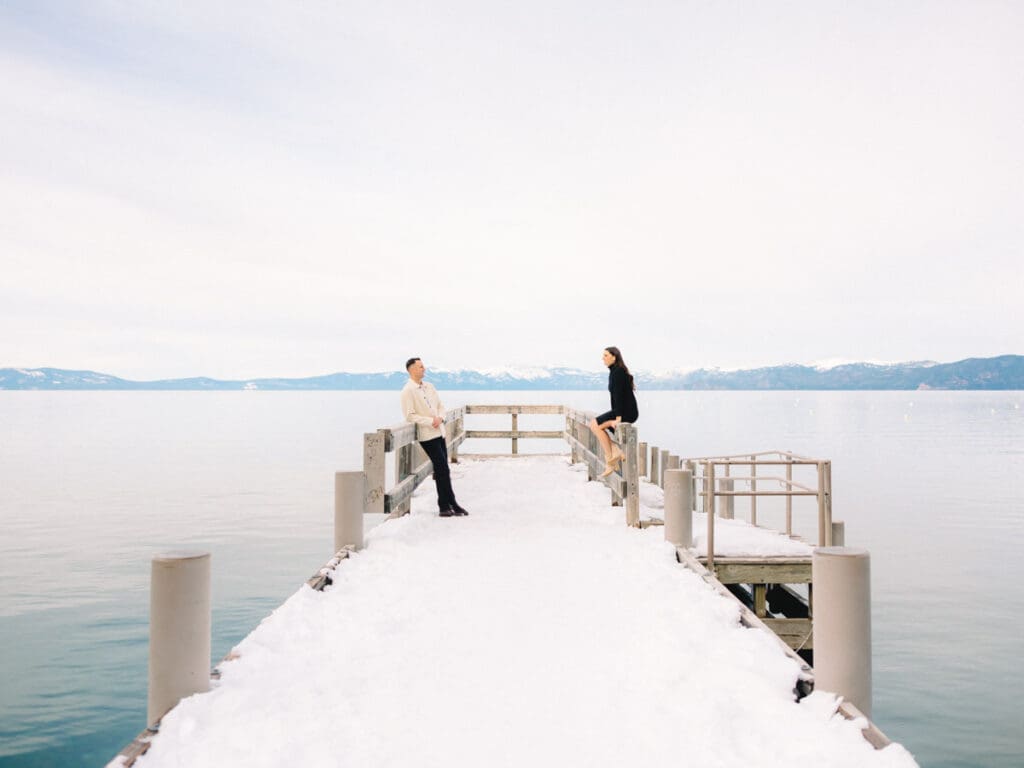 A couple stands together on a dock at Lake Tahoe, surrounded by serene water and majestic mountains in the background celebrating their engagement