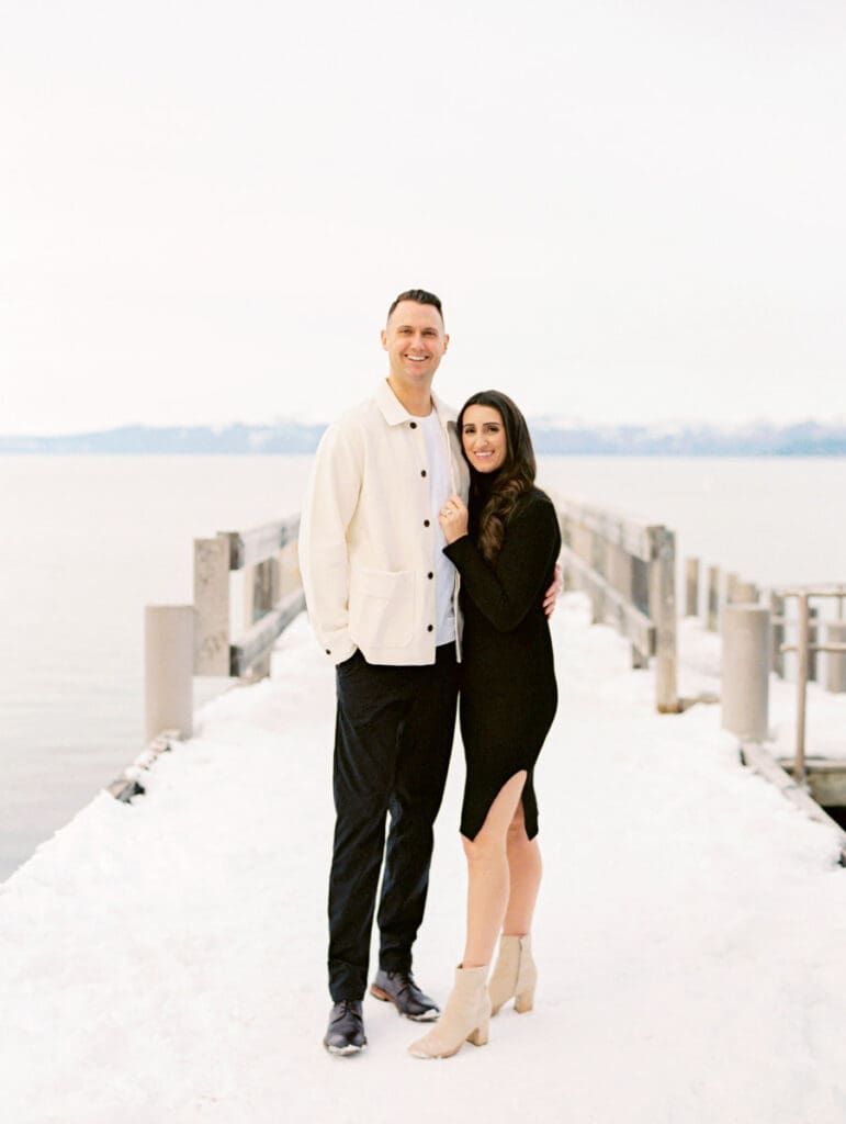 A couple stands together on a dock at Lake Tahoe, surrounded by serene water and majestic mountains in the background celebrating their engagement