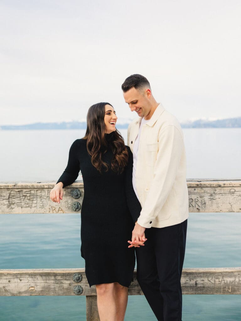 A couple stands together on a dock at Lake Tahoe, surrounded by serene water and majestic mountains in the background celebrating their engagement