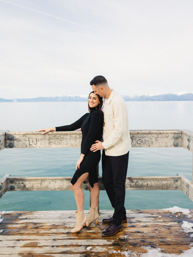 A couple stands together on a dock at Lake Tahoe, surrounded by serene water and majestic mountains in the background celebrating their engagement