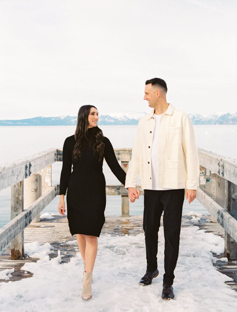 A couple stands together on a dock at Lake Tahoe, surrounded by serene water and majestic mountains in the background celebrating their engagement