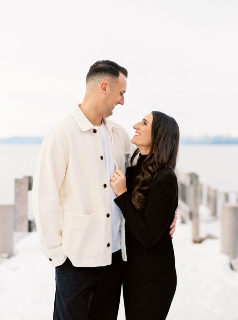 A couple stands together on a dock at Lake Tahoe, surrounded by serene water and majestic mountains in the background celebrating their engagement