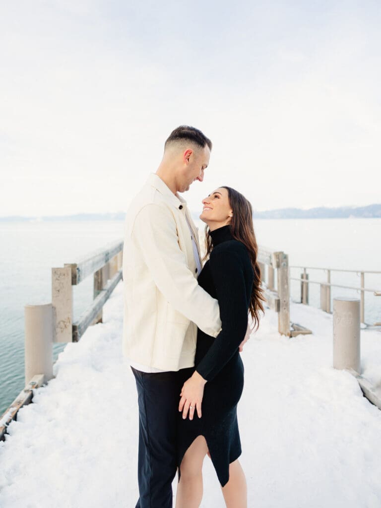 A couple stands together on a dock at Lake Tahoe, surrounded by serene water and majestic mountains in the background celebrating their engagement