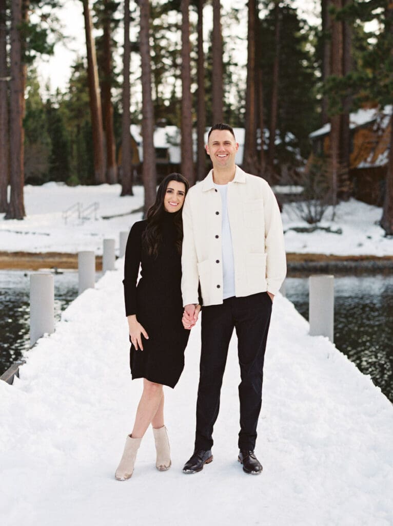 A couple stands together on a dock at Lake Tahoe, surrounded by serene water and majestic pine trees in the background celebrating their engagement