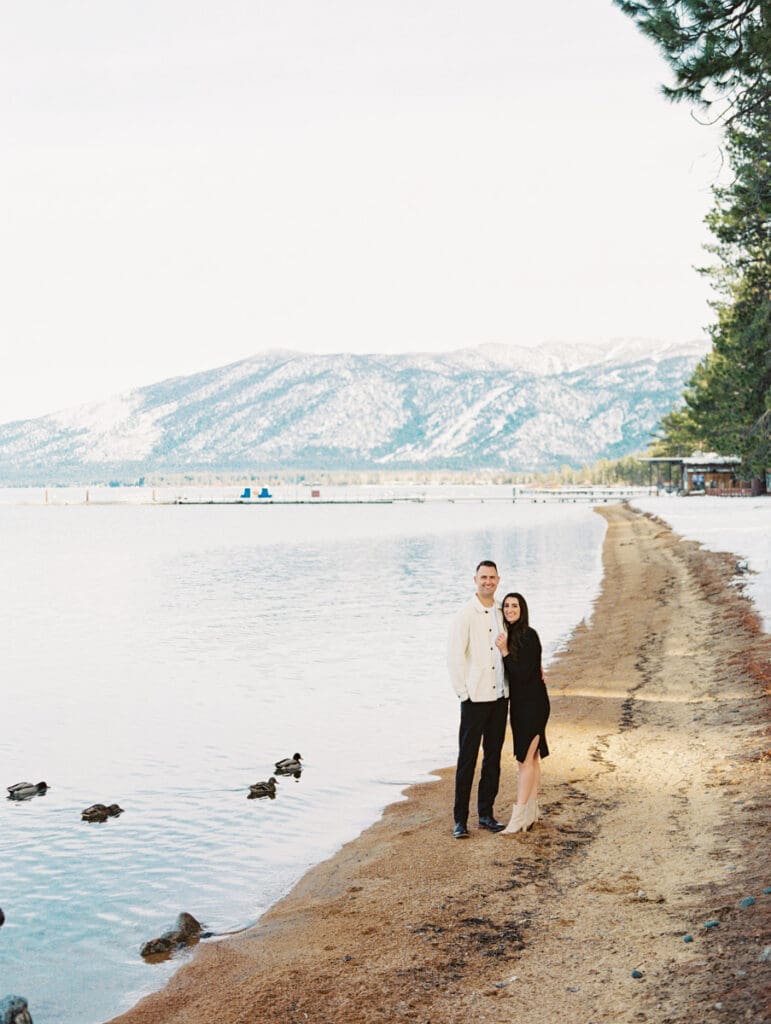 A couple stands together on the beach in Lake Tahoe, surrounded by serene water and majestic mountains in the background celebrating their engagement