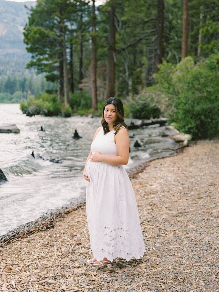 A pregnant woman in a white dress stands gracefully by the water during a maternity photography session in South Lake Tahoe.