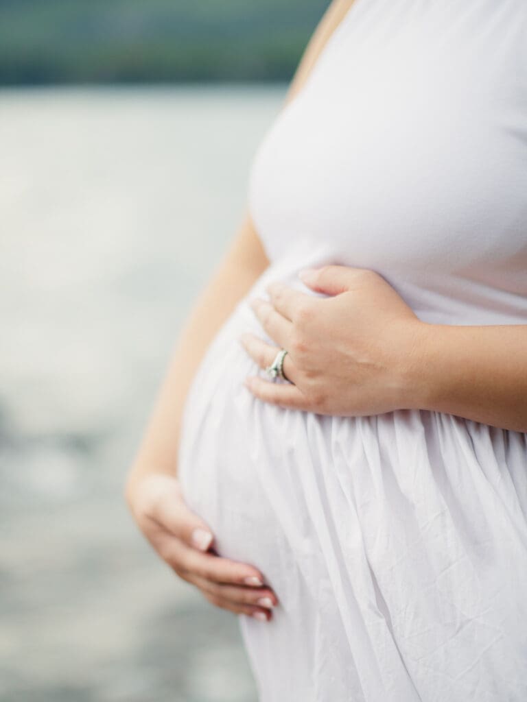 A pregnant woman in a white dress stands gracefully by the water during a maternity photography session in South Lake Tahoe.