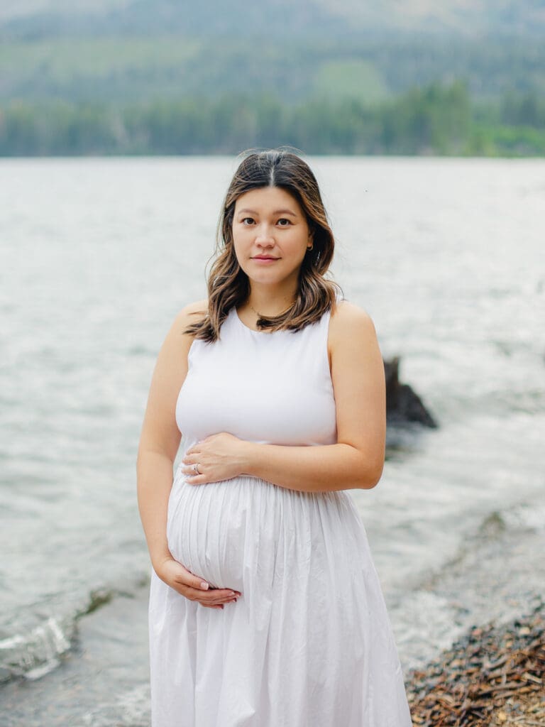 A pregnant woman in a white dress stands gracefully by the water during a maternity photography session in South Lake Tahoe.