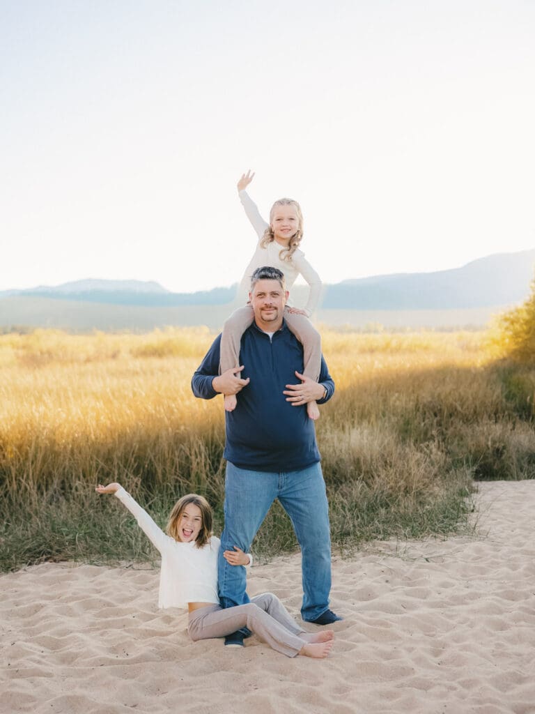 A family photo session at South Lake Tahoe features a father and his two girls in white dresses standing on the sandy shore.