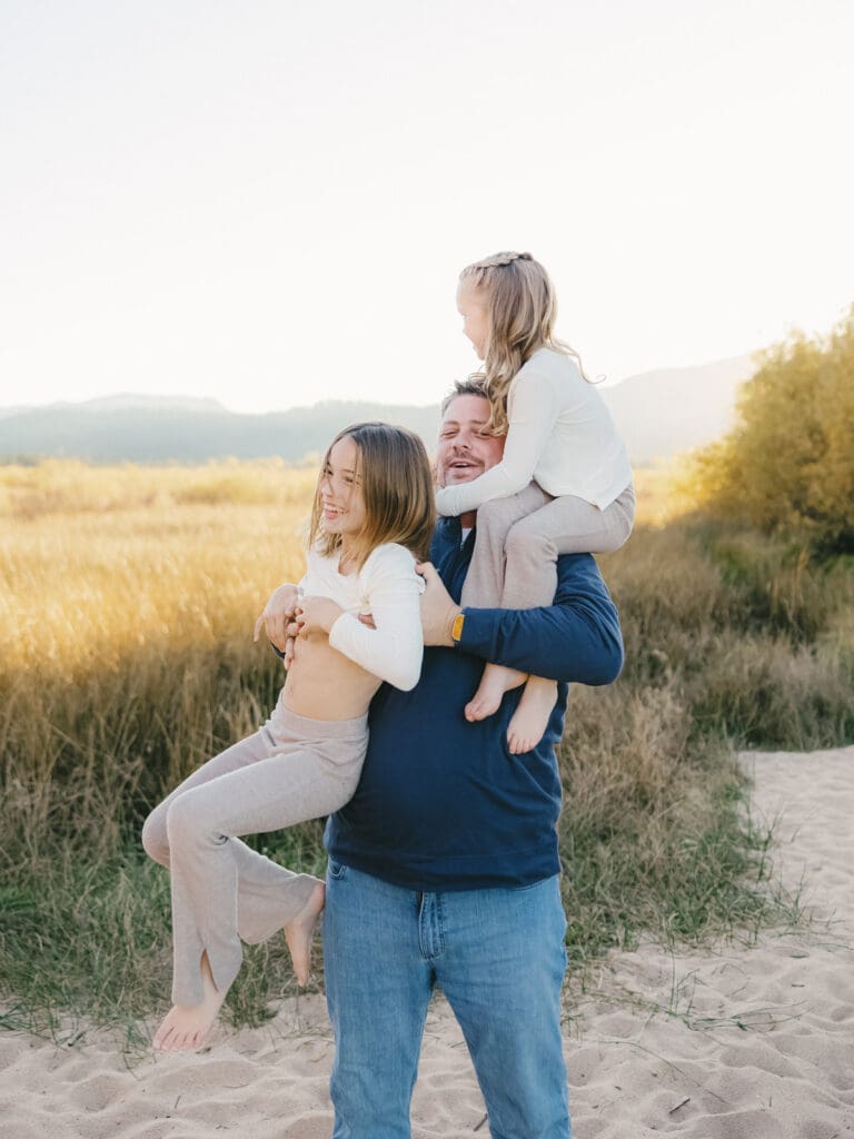 A family photo session at South Lake Tahoe features a father and his two girls in white dresses standing on the sandy shore.