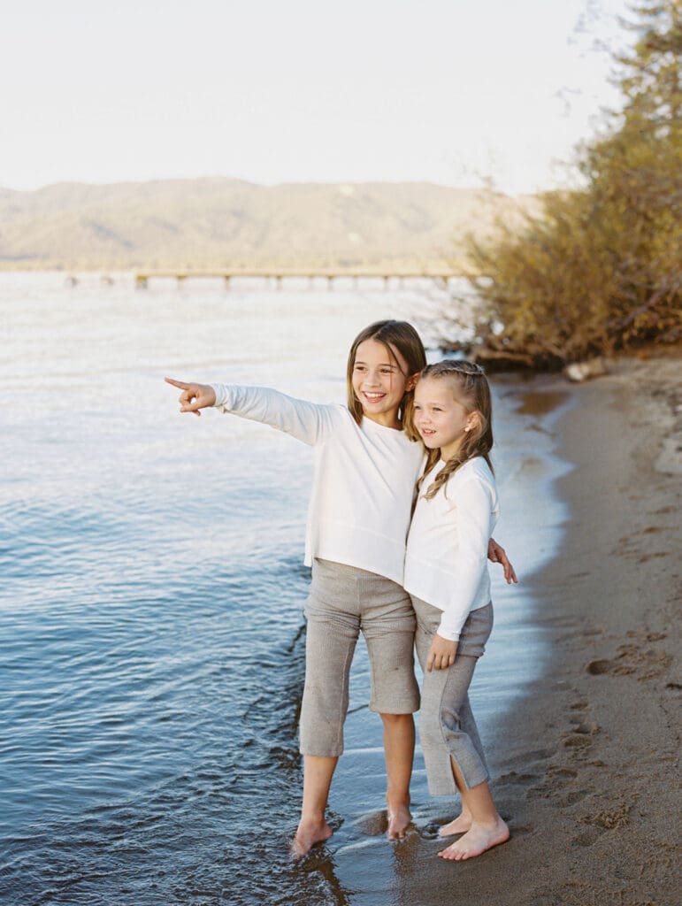 Two little girls dressed in white shirts stand on the beach, part of a family photo session in South Lake Tahoe.