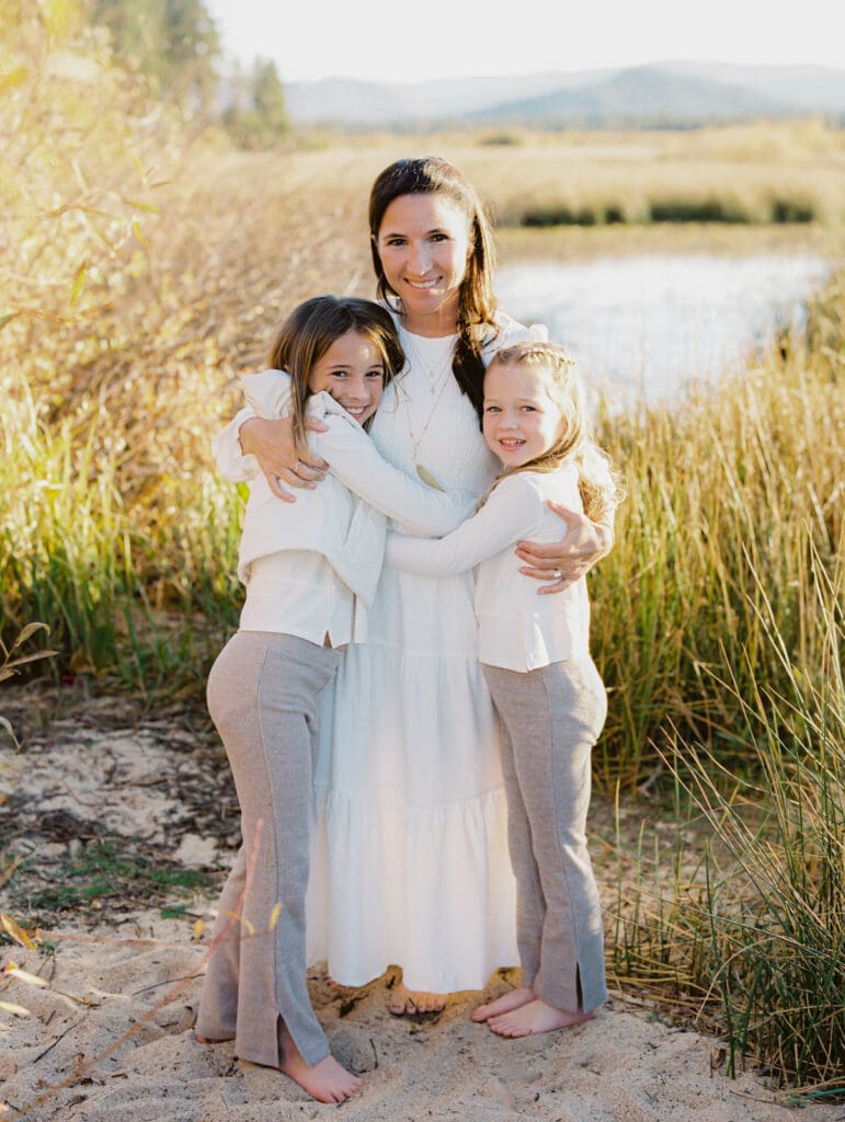 A family photo session at South Lake Tahoe features a mother and her two girls in white dresses standing on the sandy shore.