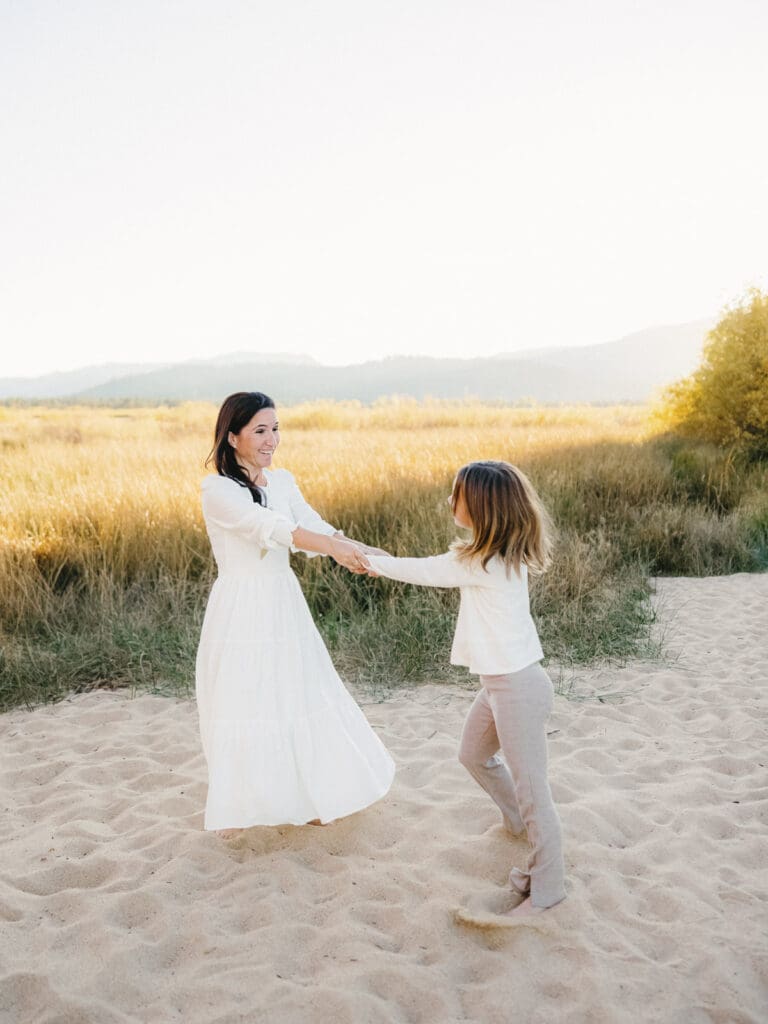 A family photo session at South Lake Tahoe features a mother and her daughter in white dresses standing on the sandy shore.