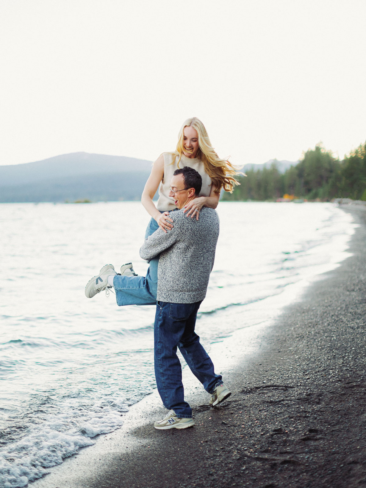 A couple enjoys an engagement session at Lake Tahoe, with the man playfully carrying the woman on his back at the beach.